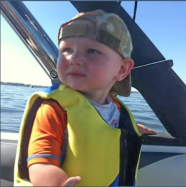 A young child wearing a life jacket and a cap, looking out at the water while on a boat.