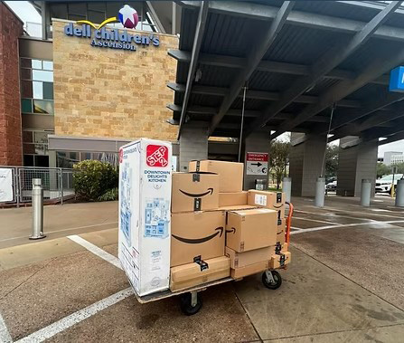 A cart loaded with boxes, including a large item labeled "Step2" and several smaller cardboard boxes, is positioned outside Dell Children's Medical Center.
