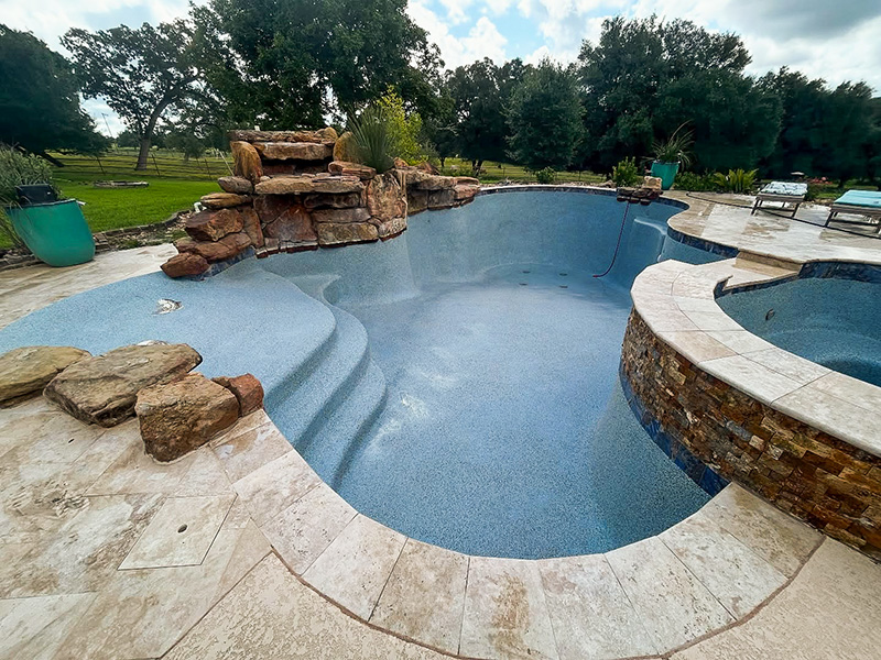 A partially empty swimming pool with a textured blue surface, natural stone accents, and surrounding patio area. Trees and grass are visible in the background.