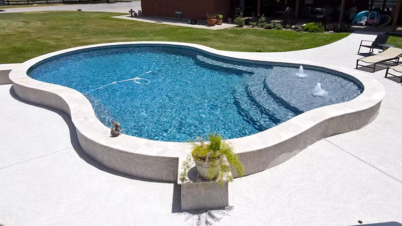 freeform-pools A large, kidney-shaped swimming pool with clear blue water, a gentle slope at one end, and two small fountains, surrounded by a concrete deck and a landscaped area with a potted plant.