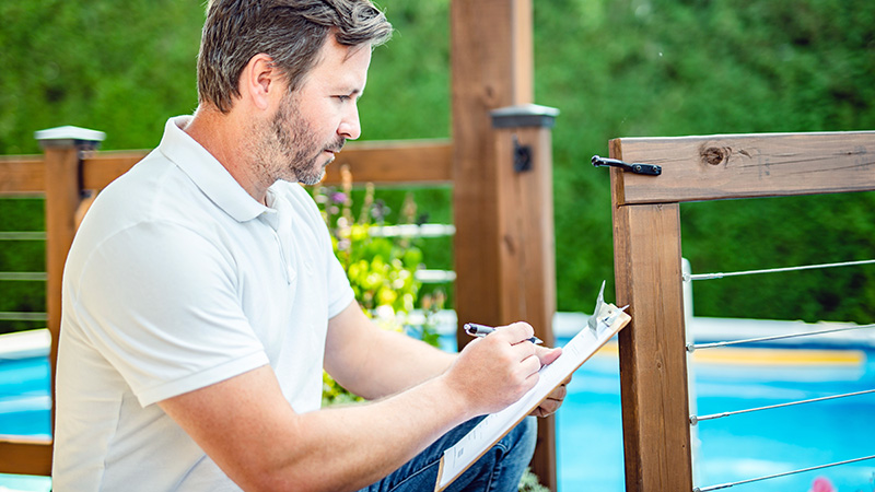 A man sitting near a pool is writing notes on a clipboard while reviewing a wooden fence.
