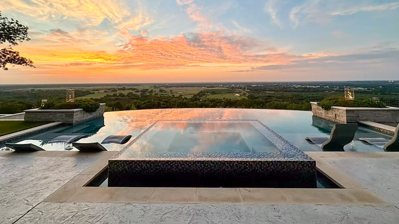negative-edge-pool A luxurious infinity pool overlooking a sunset-lit landscape with clouds and rolling hills in the distance.