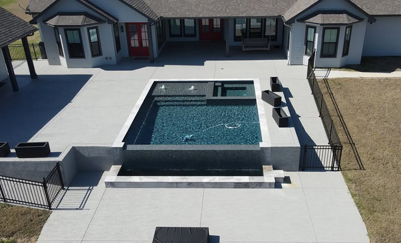 new-pool-construction-gallery-03 A modern, rectangular swimming pool with a water feature, surrounded by concrete decking and a grass area, adjacent to a house with large windows and a black fence.