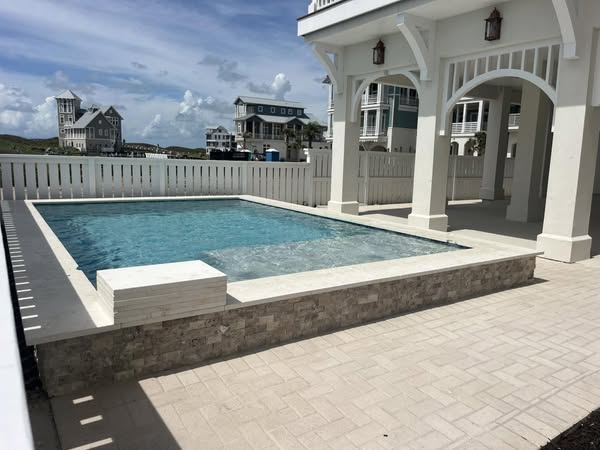 new-pool-construction-gallery-04 A rectangular swimming pool surrounded by a paved area and white railing, with houses in the background under a blue sky with clouds.