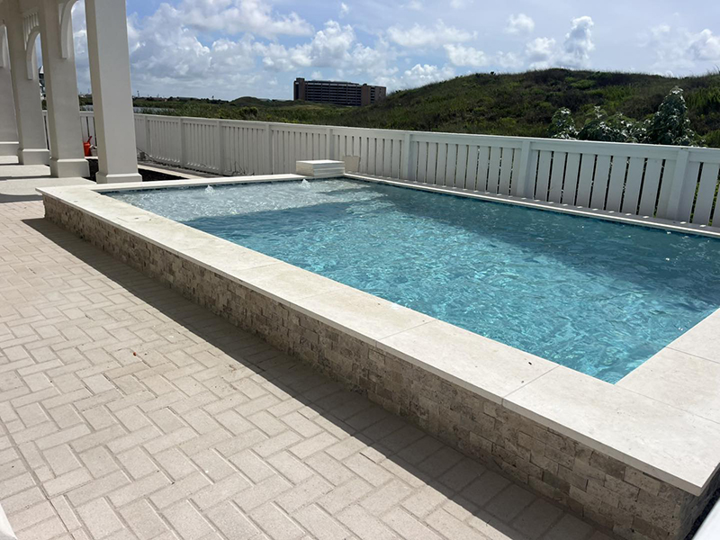 new-pool-construction-gallery-06 A clear swimming pool with a stone border, surrounded by a patio area and a white railing, set against a green landscape and blue sky.