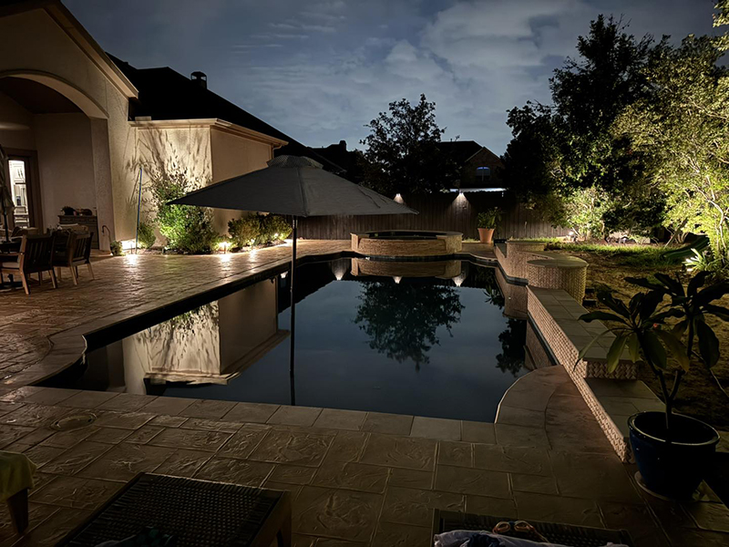 new-pool-construction-gallery-07 A serene backyard at night featuring a swimming pool with reflections, illuminated landscaping, and a cantilevered umbrella beside the pool.