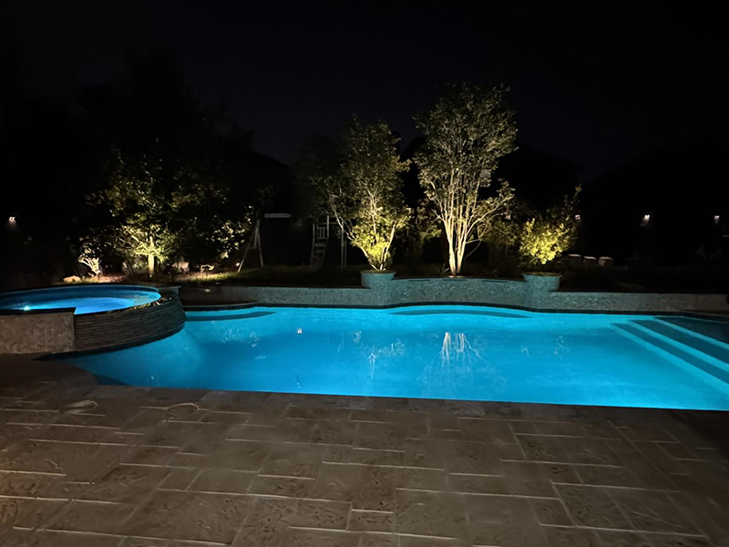 new-pool-construction-gallery-08 A softly lit swimming pool at night, surrounded by trees and illuminated landscaping.