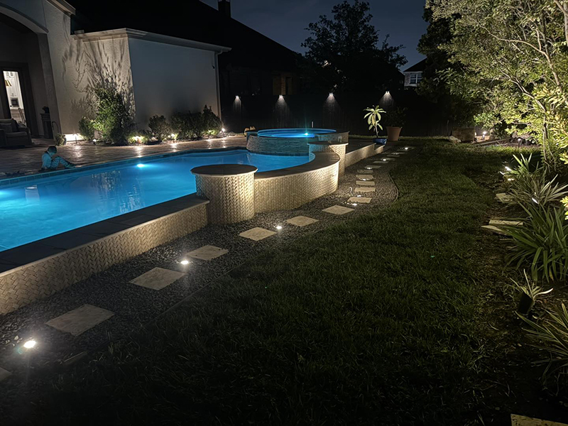 new-pool-construction-gallery-09 A night view of a landscaped backyard featuring a blue-lit swimming pool, a spa, and illuminated pathway stones surrounded by grass and plants.