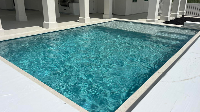 A clear swimming pool filled with blue water, surrounded by a light-colored deck and columns.