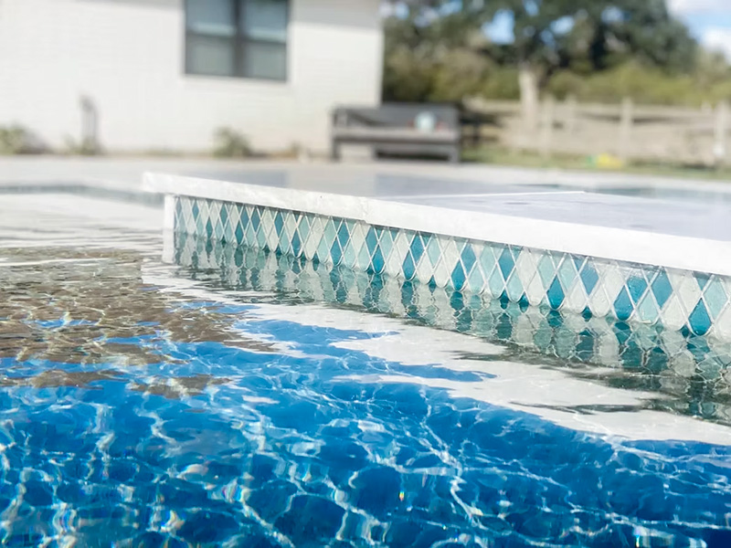 A close-up view of a swimming pool's edge with a decorative tiled border, reflecting the clear blue water and a background of a house and bench.