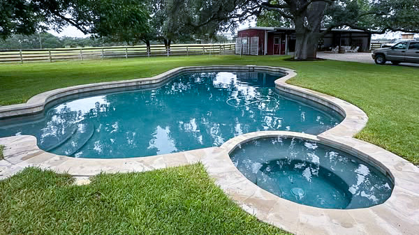 A pool shaped like an abstract figure, surrounded by grass and trees, with a small attached spa. In the background, there is a barn and a parked vehicle.