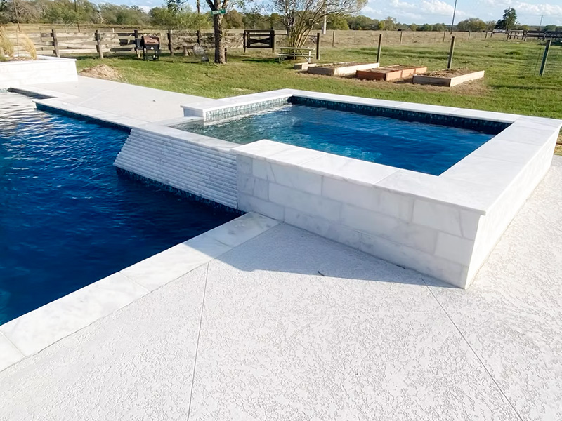 A modern pool area with a blue water pool and a spa, featuring white stone edges and a textured concrete deck. The background shows a grassy landscape with trees and a fence.