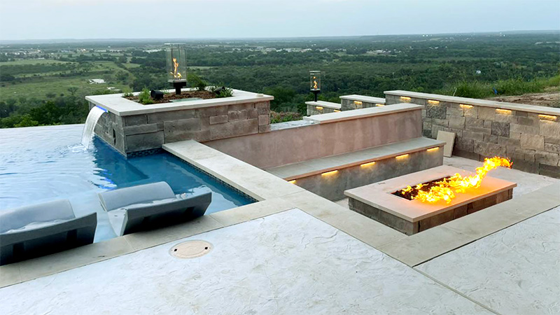 A luxurious outdoor area featuring an infinity pool, a fire pit with flames, and scenic views of a green landscape.