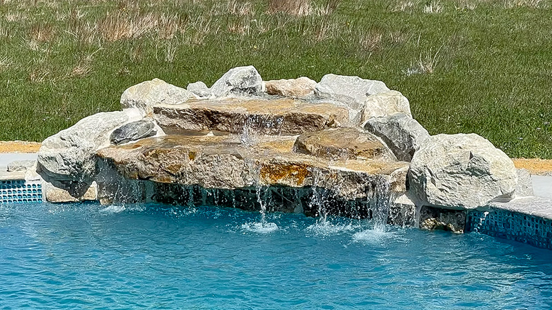 A stone waterfall feature over a swimming pool, with clear blue water and a grassy area in the background.