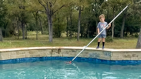 A child stands by a swimming pool, using a pole to clean the water. Surrounding trees are visible in the background.