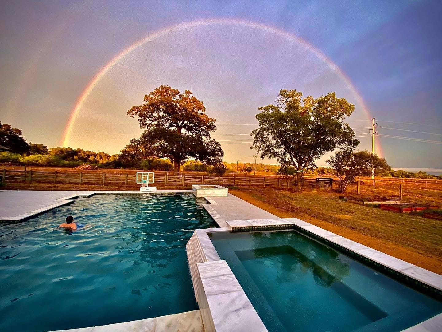 Custom Built Pools A custom built swimming pool with a spa and a person swimming in it under a rainbow.
